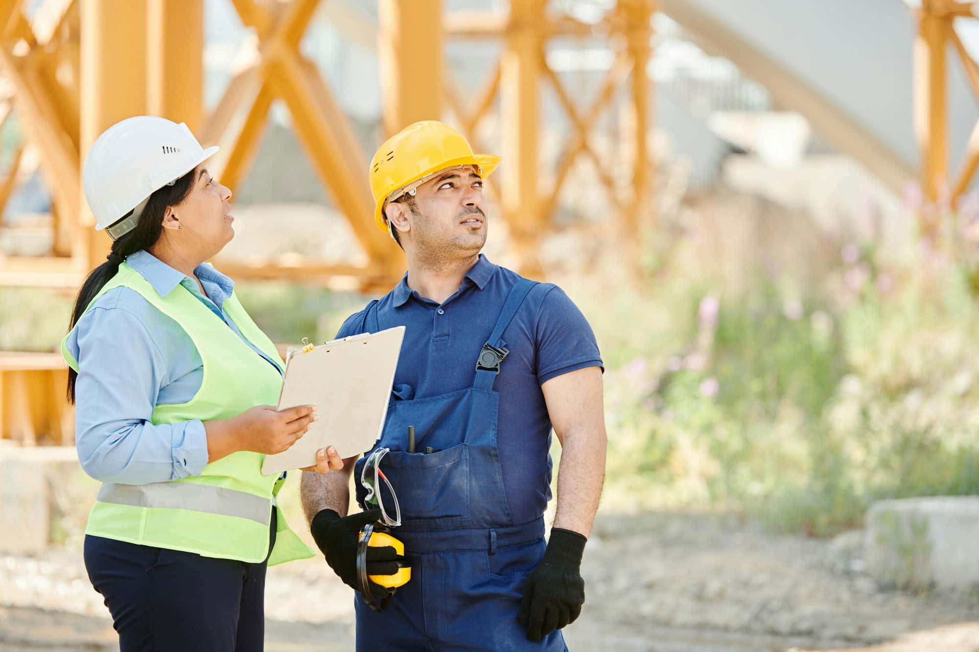 Bauleiterin und Bauarbeiter mit Schutzhelmen und Klemmbrett auf einer Baustelle im Gespräch.