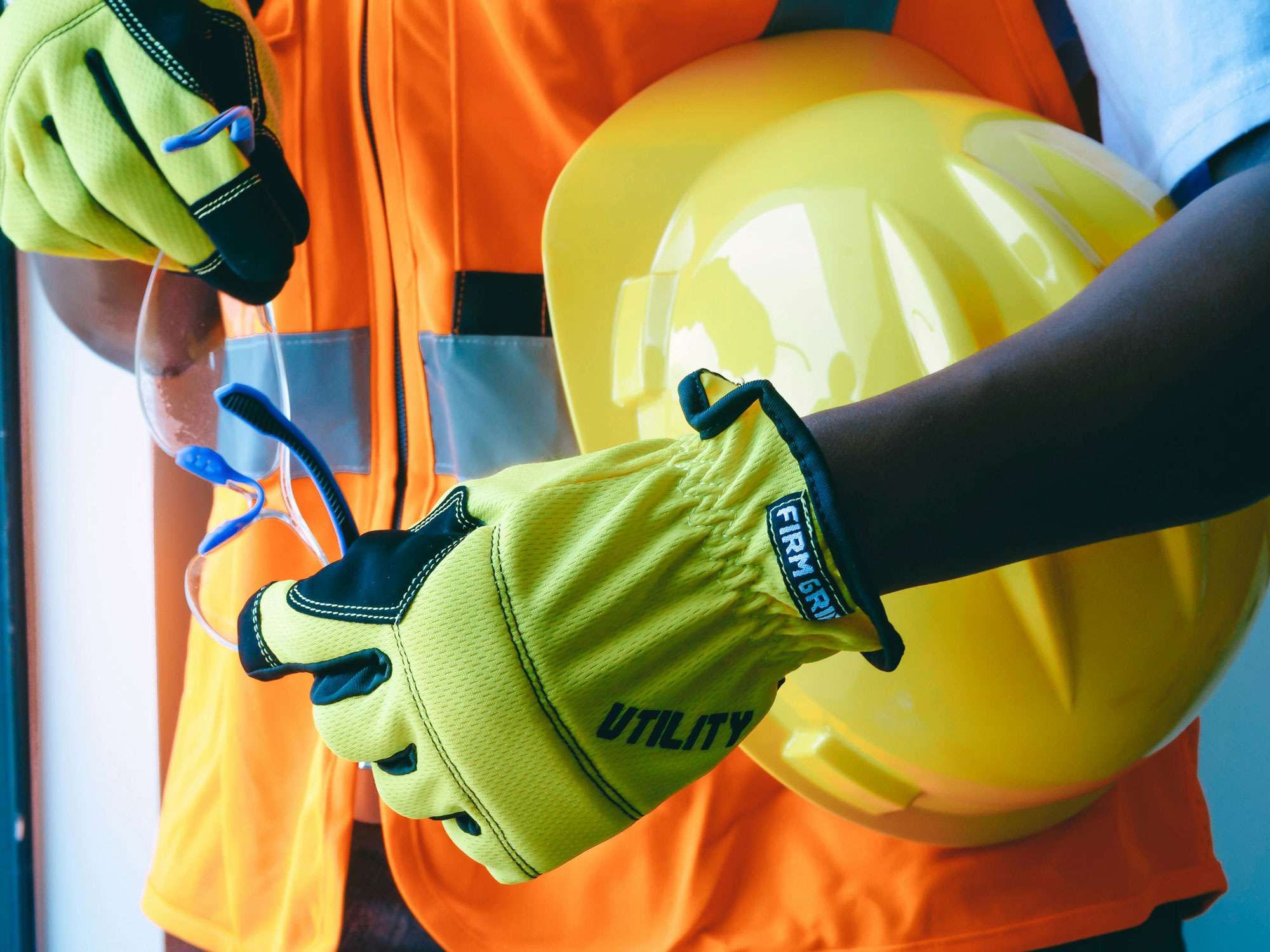 Person mit gelben Schutzhandschuhen, Schutzbrille und gelbem Schutzhelm in orangefarbener Warnweste.
