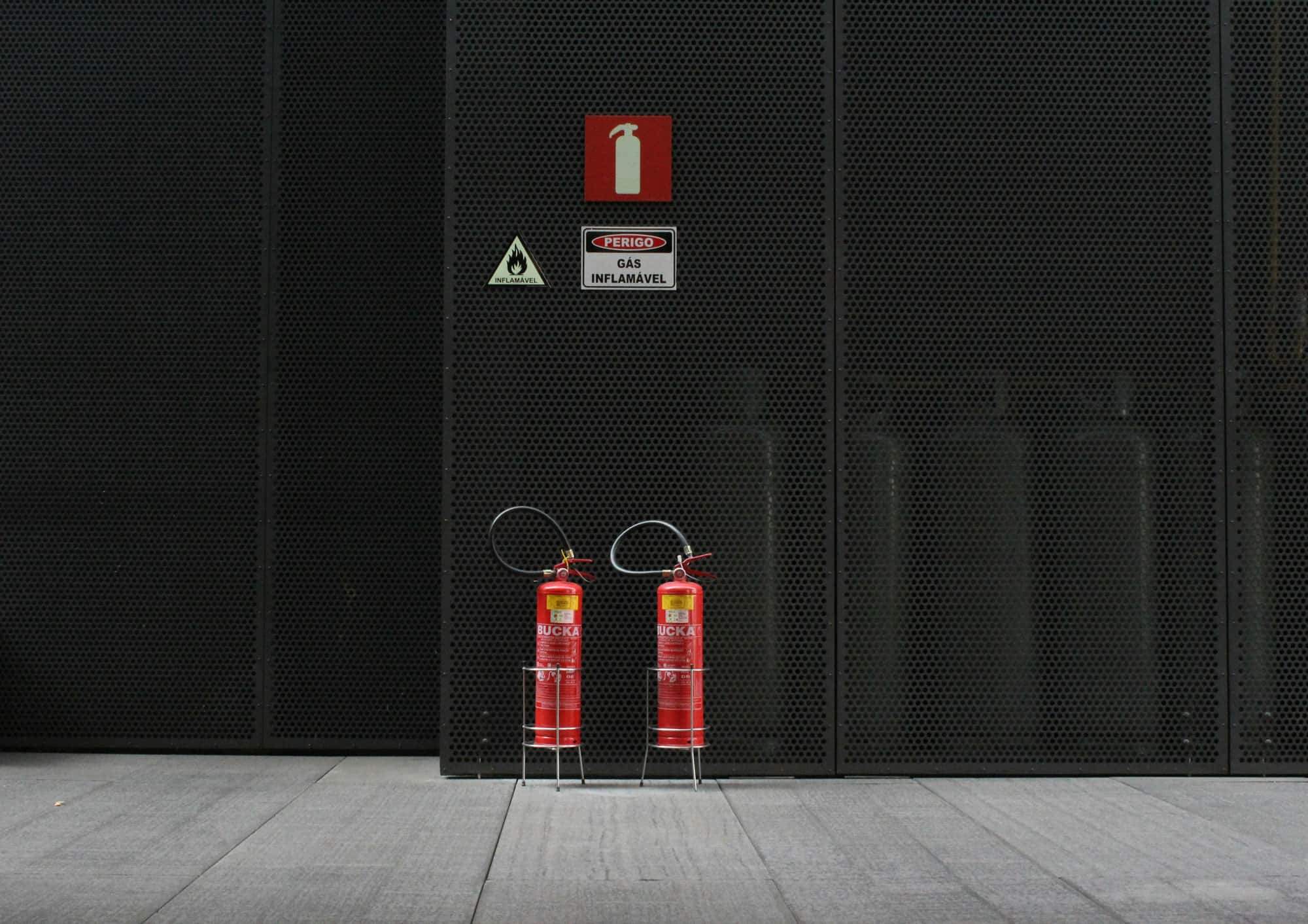 Zwei Feuerlöscher vor einer dunklen Metallwand mit Hinweisschildern für Brandgefahr und entflammbares Gas.