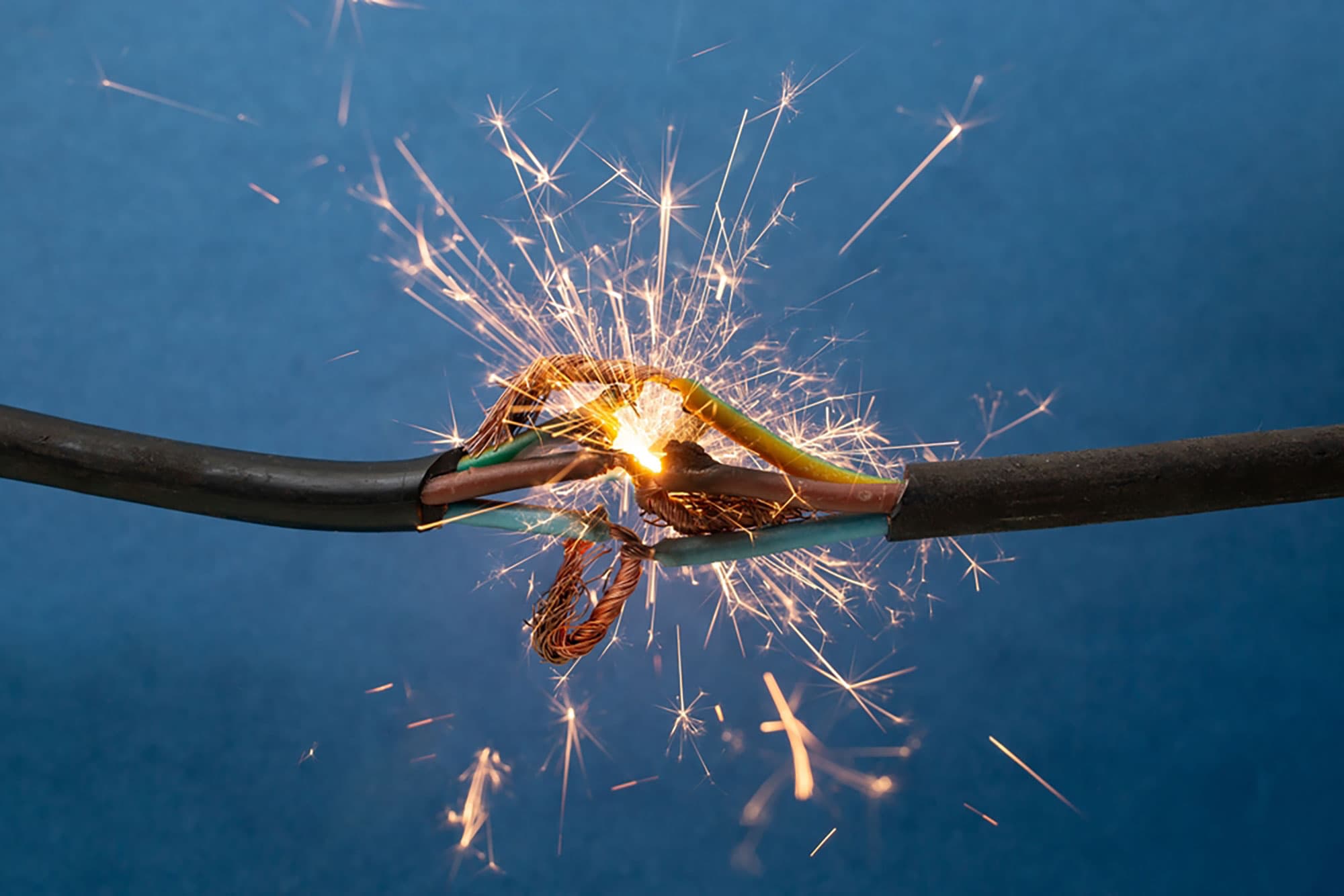 Ein defektes Stromkabel mit freiliegenden Drähten verursacht Funken und einen Lichtblitz vor blauem Hintergrund.