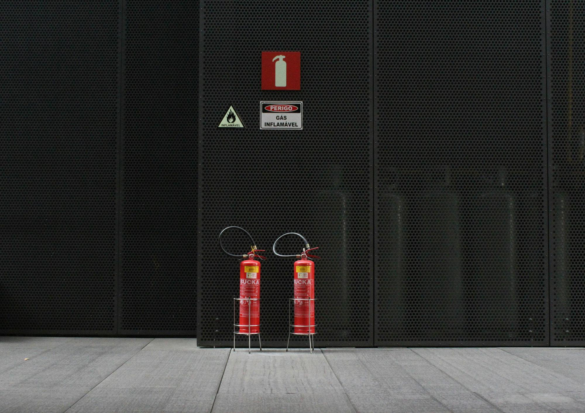 Zwei Feuerlöscher vor einer dunklen Metallwand mit Hinweisschildern für Brandgefahr und entflammbares Gas.