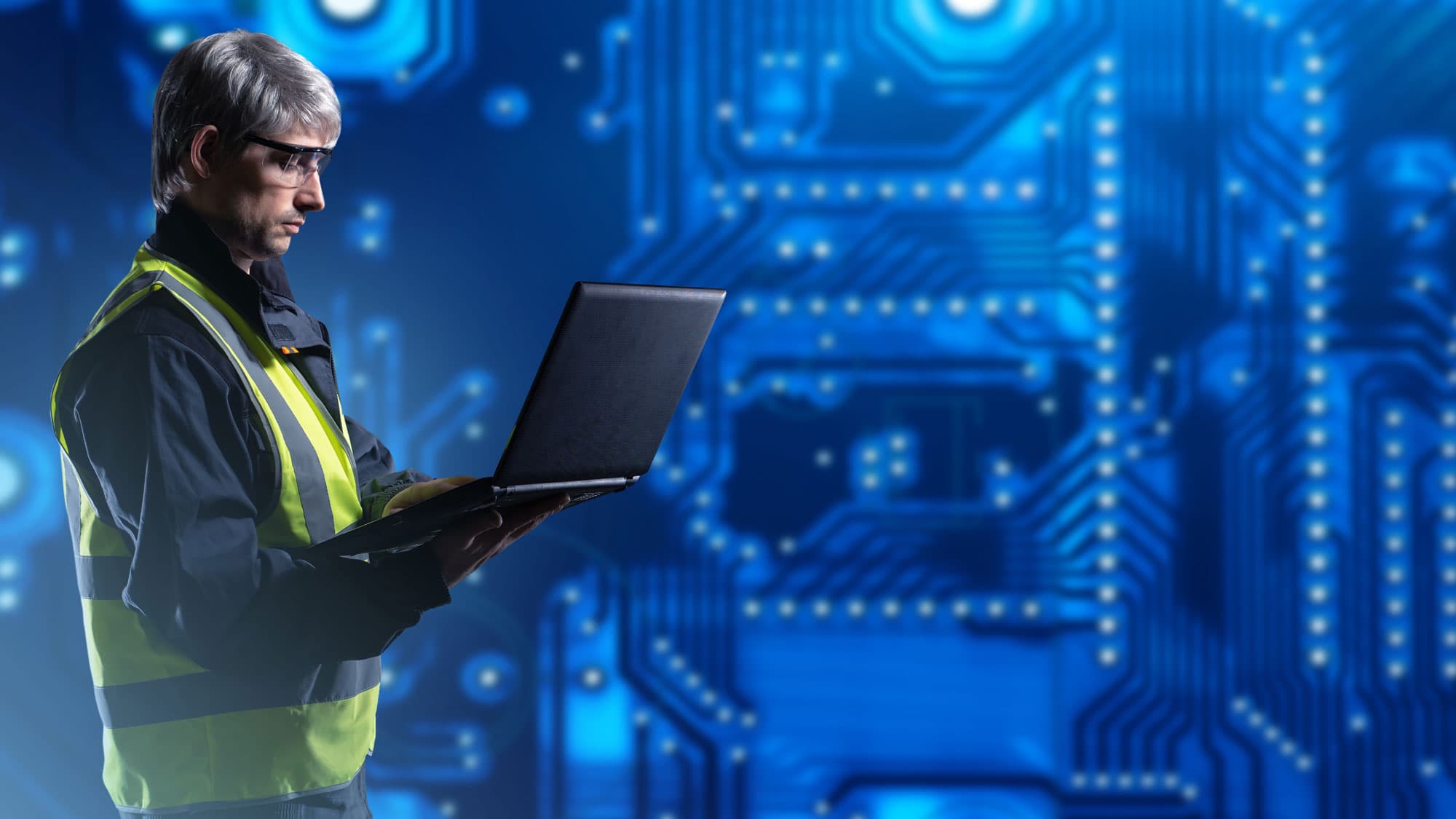 Ein Techniker in Sicherheitsweste und Schutzbrille arbeitet an einem Laptop vor einem digitalen, blau leuchtenden Platinen-Hintergrund.