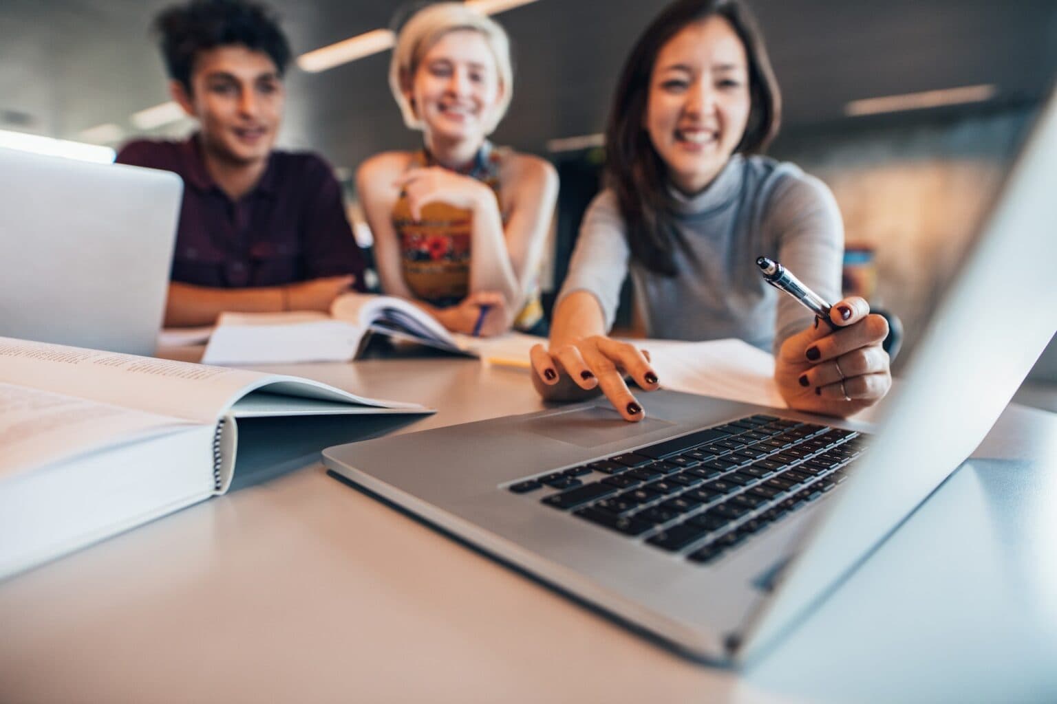 Drei Personen sitzen an einem Tisch und arbeiten gemeinsam an einem Laptop. Sie lachen und diskutieren, während sie auf den Bildschirm schauen. Bücher und Notizblöcke liegen auf dem Tisch, was auf eine gemeinsame Lern- oder Arbeitsumgebung hinweist.